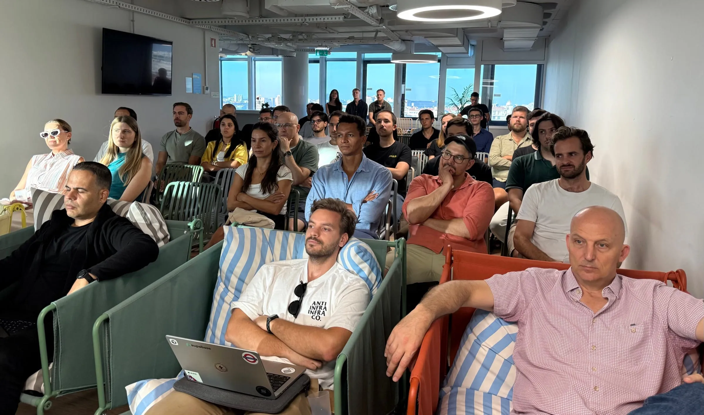 A full room of attendees on striped benches, watching a talk, with the Lisbon skyline through the windows behind them