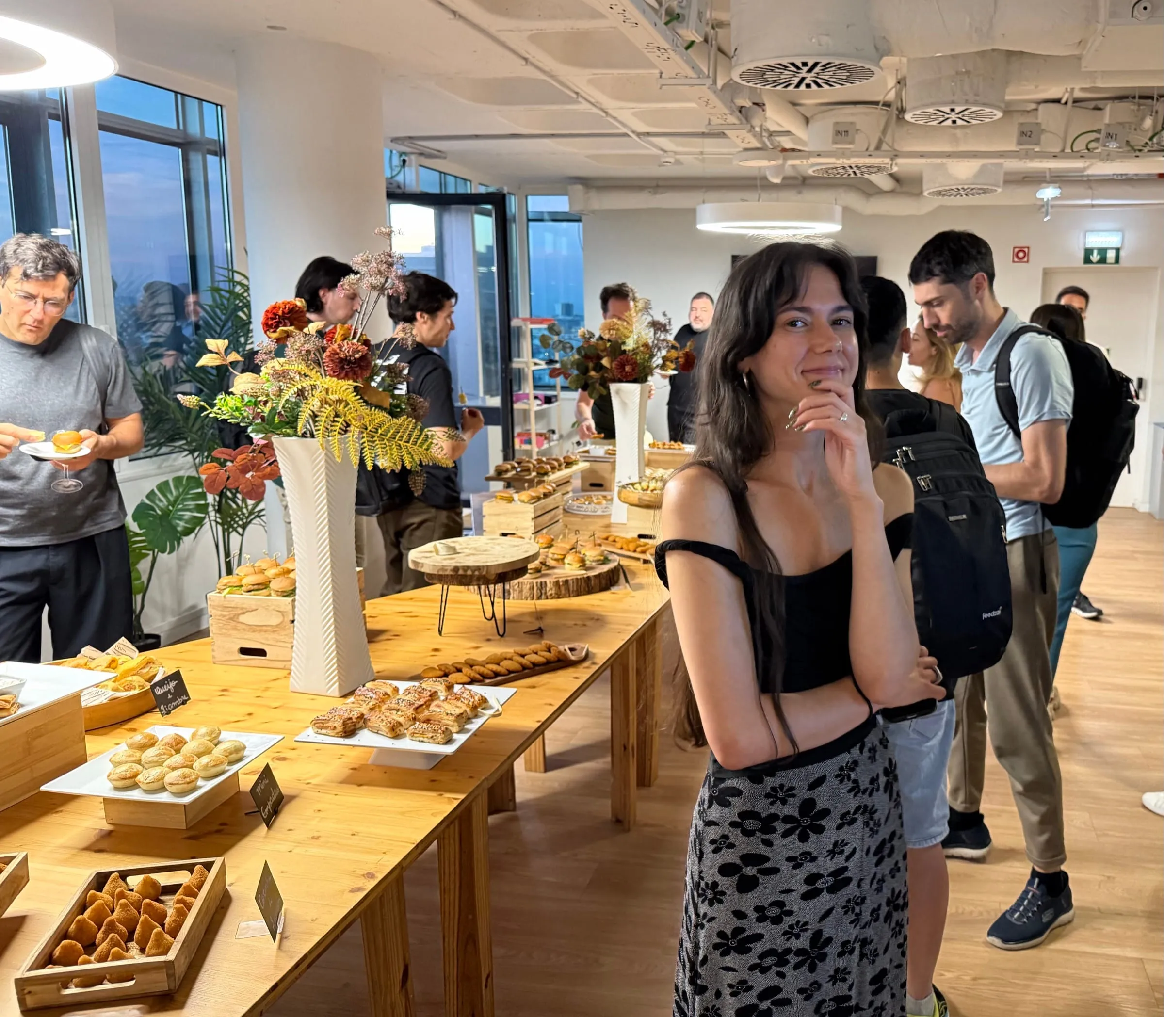 A guest smiles at the camera beside long wooden tables of Portuguese pastries and flowers, as others help themselves