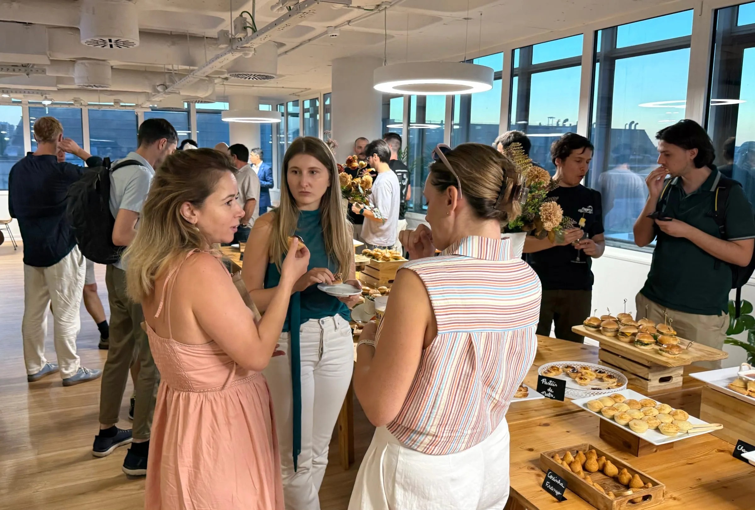 Three guests chat over plates of snacks by the buffet, with evening light coming through the windows