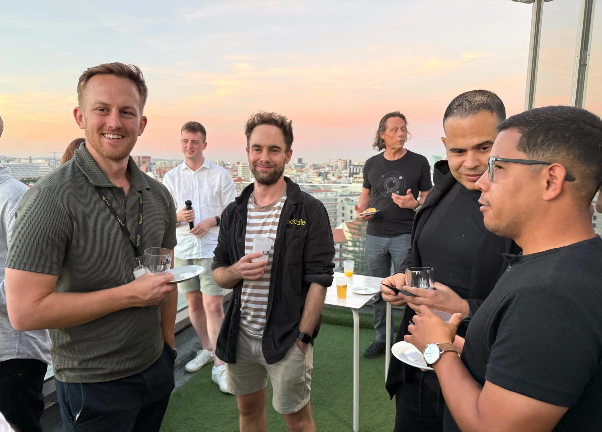 Attendees, two in Paddle-branded clothing, chat over drinks on a rooftop terrace as the sun sets over Lisbon